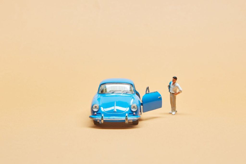 A miniature figurine stands beside a classic blue toy car on a beige background, creating a whimsical scene.